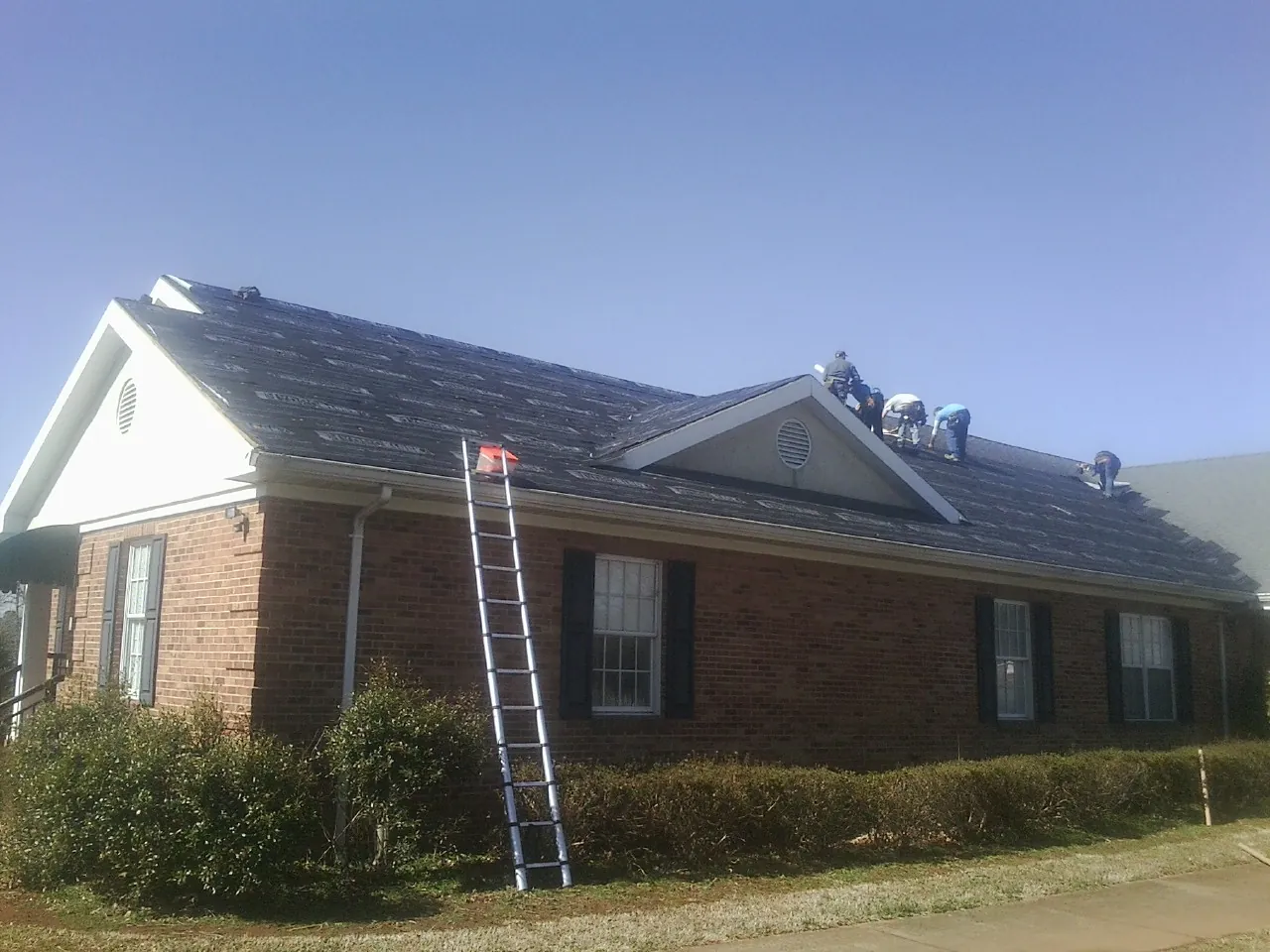 Crew tearing shingles off a brick ranch roof with a ladder set — Metal Roof Repair work by our crew in Gunter