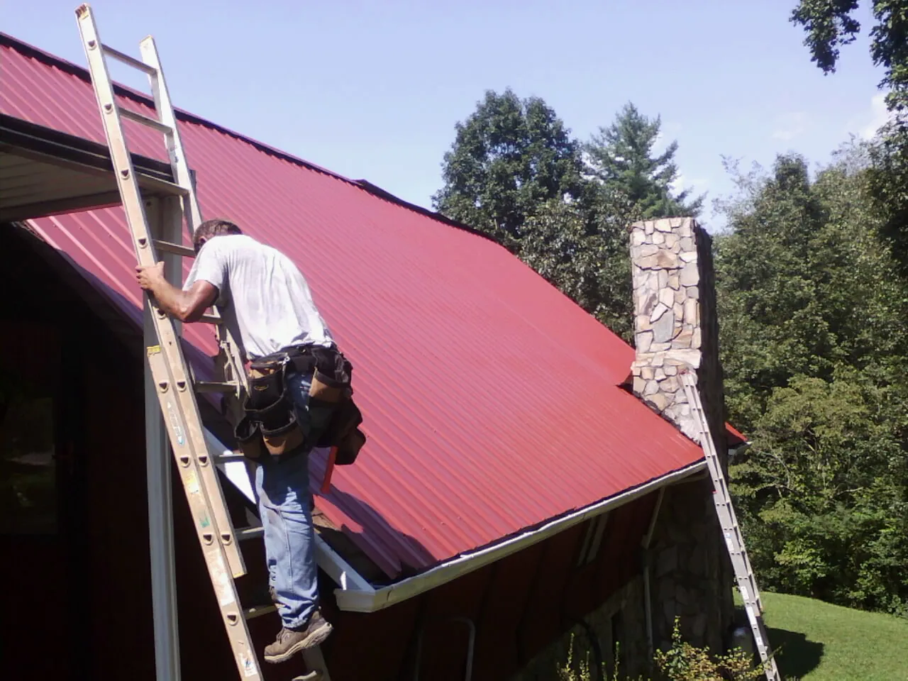 Roofer on a ladder beside a red metal roof and stone chimney — Soffit & Fascia Repair by Roofing Brothers in Gunter